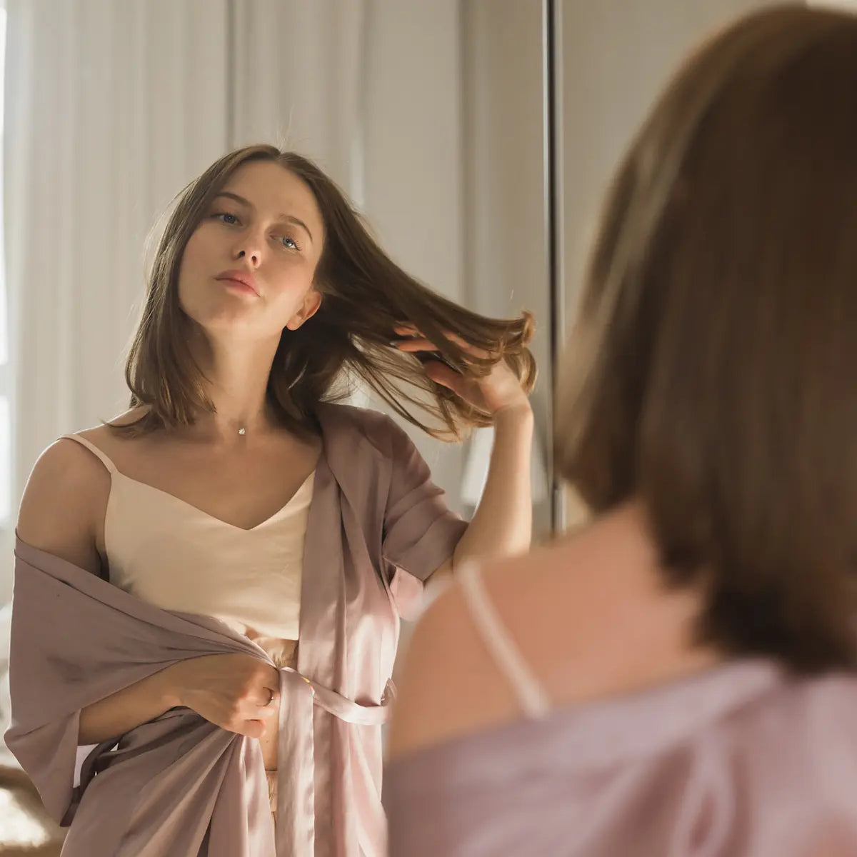 Woman in silk robe inspecting her medium-length brown hair in the mirror before a haircut