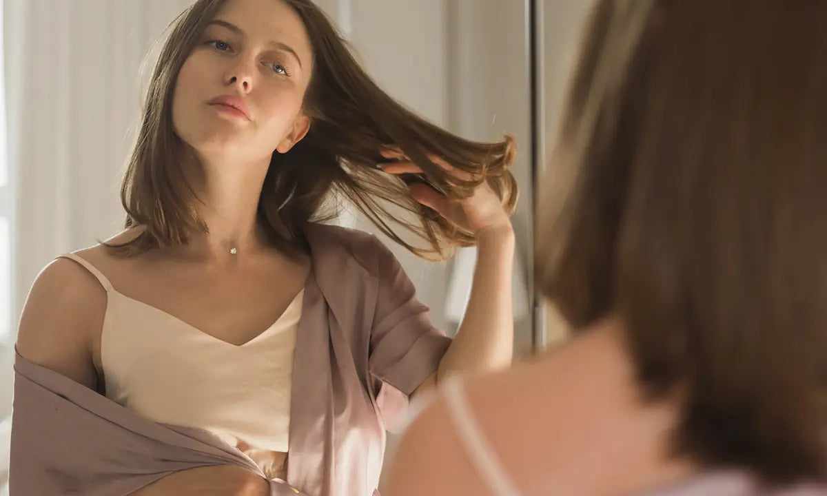 Woman in silk robe inspecting her medium-length brown hair in the mirror before a haircut